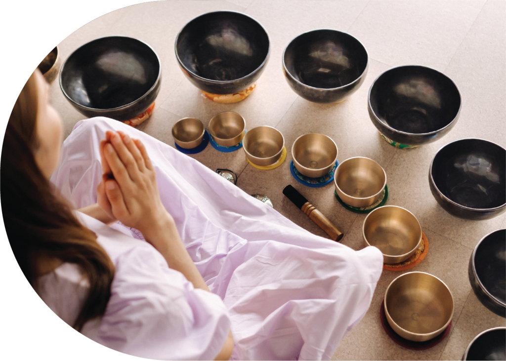 A woman in a white dress sits cross-legged with hands in prayer position, surrounded by various metal singing bowls and a mallet arranged on the floor, highlighting some of the Additional Offerings available during her tranquil session.