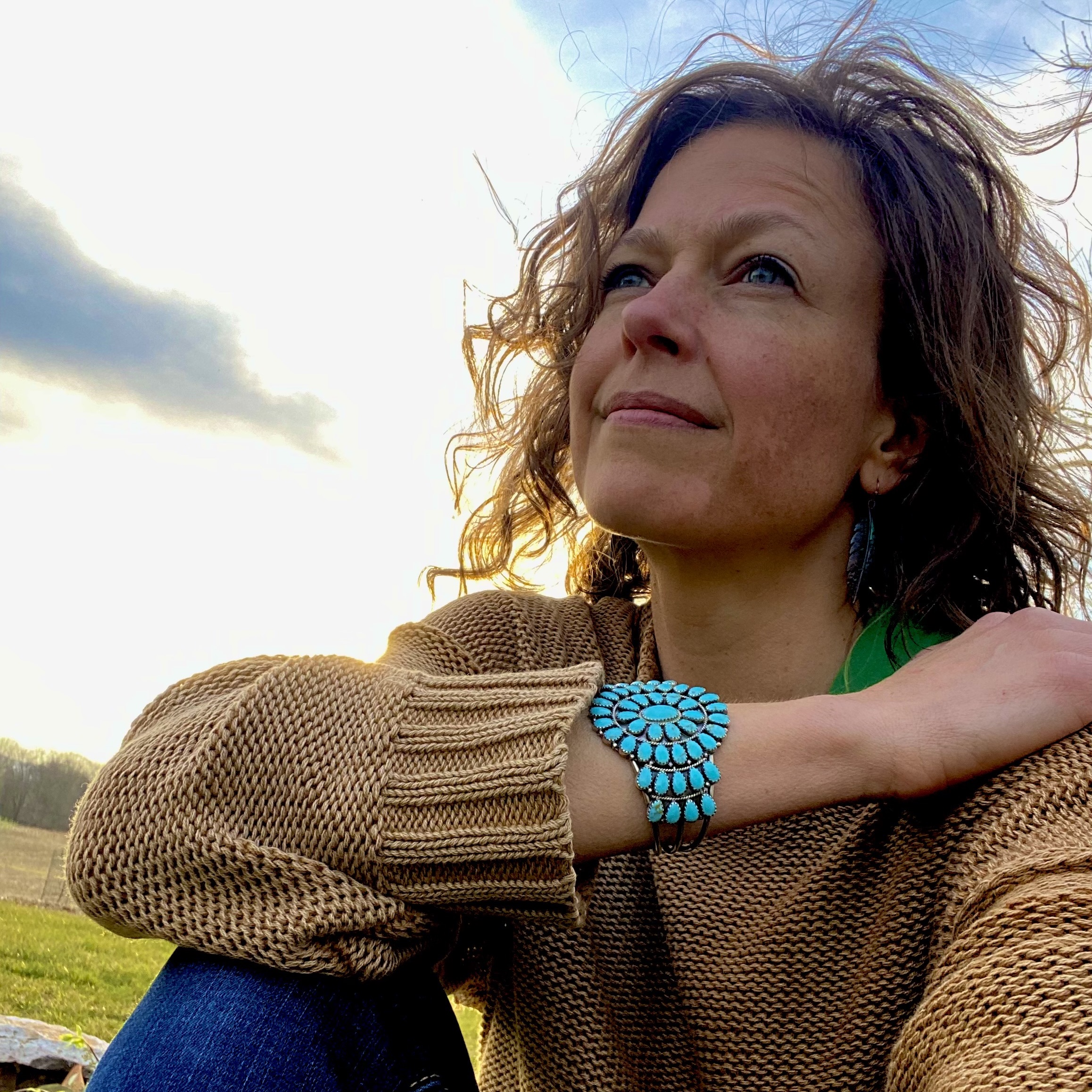 Annette, a Spiritual Director with wavy brown hair, sits outdoors on the grass in a tan sweater and blue jeans, wearing a large turquoise bracelet and gazing thoughtfully toward the sky on a partly cloudy day.