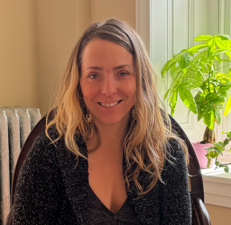 A woman with long, wavy blonde hair smiles while sitting indoors near a window. The massage therapist wears a dark textured sweater, and a green plant is visible in the sunlight on the windowsill behind her.