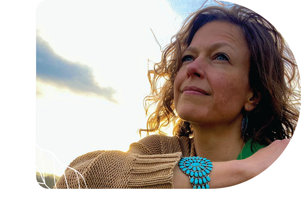 Spiritual Director Annette, with curly hair, gazes thoughtfully into the distance at sunset, wearing a tan sweater and turquoise bracelet, framed by a faint leaf design in the background.