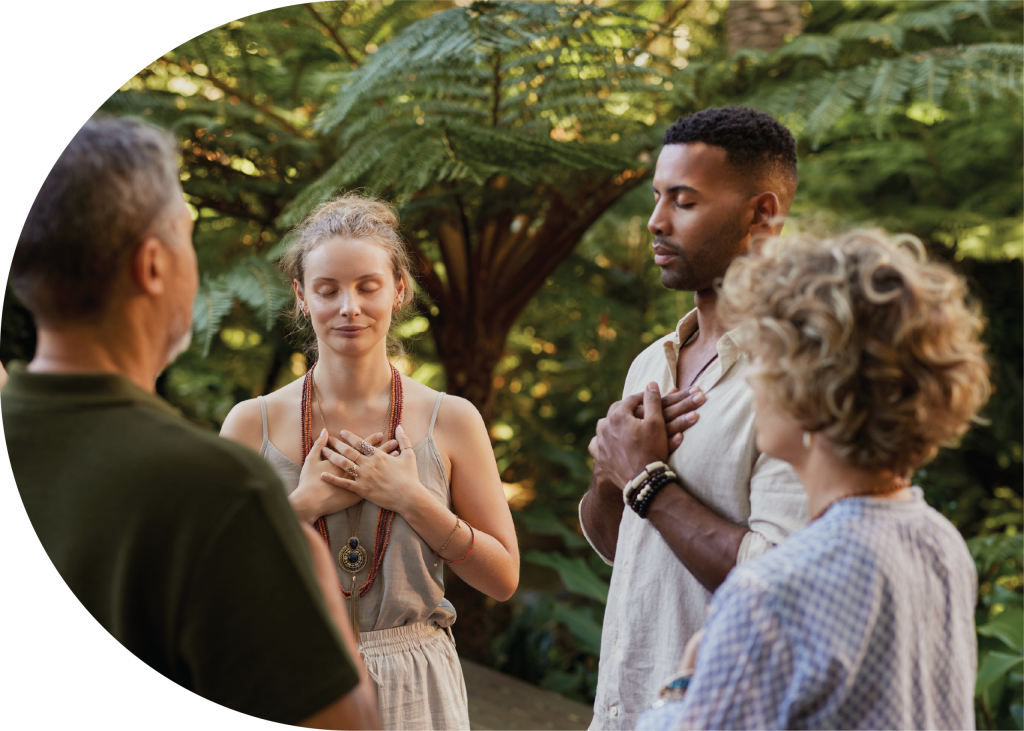 Four adults stand in a circle outdoors with eyes closed and hands on their chests, appearing calm and meditative as if guided by a spiritual companion amidst lush greenery.