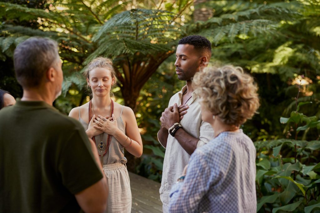 Four people stand in a lush garden, eyes closed with hands on their chests, appearing calm and peaceful as they share a mindfulness exercise—a moment of connection with themselves and each other as spiritual companions.