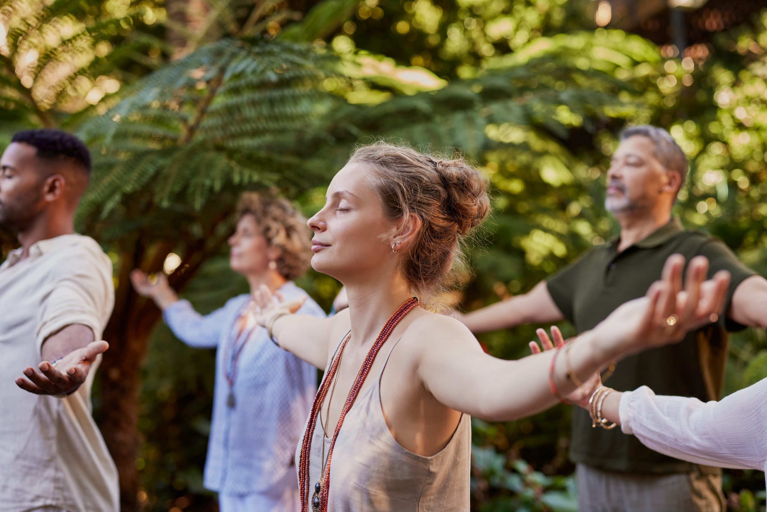 Four adults stand outdoors with eyes closed and arms outstretched, appearing relaxed and peaceful. They are surrounded by lush green foliage, suggesting a calm environment ideal for reflection or spiritual direction.